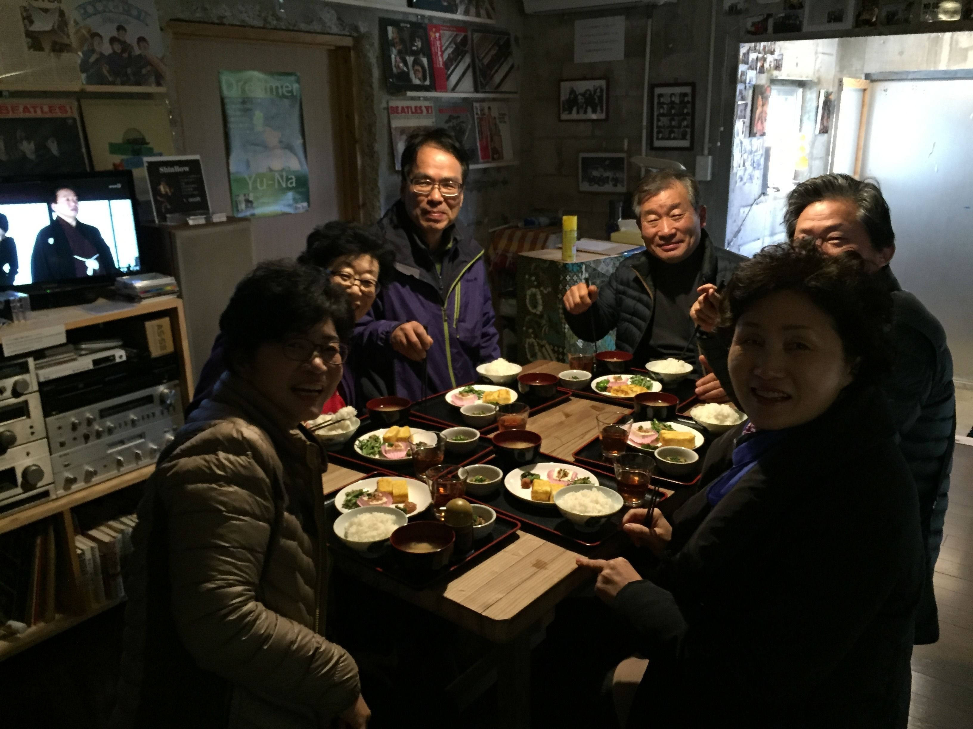 A group of people seated around a table with various dishes, in a room with posters on the walls and a television in the background.