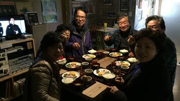 A group of people seated around a table with various dishes, in a room with posters on the walls and a television in the background.