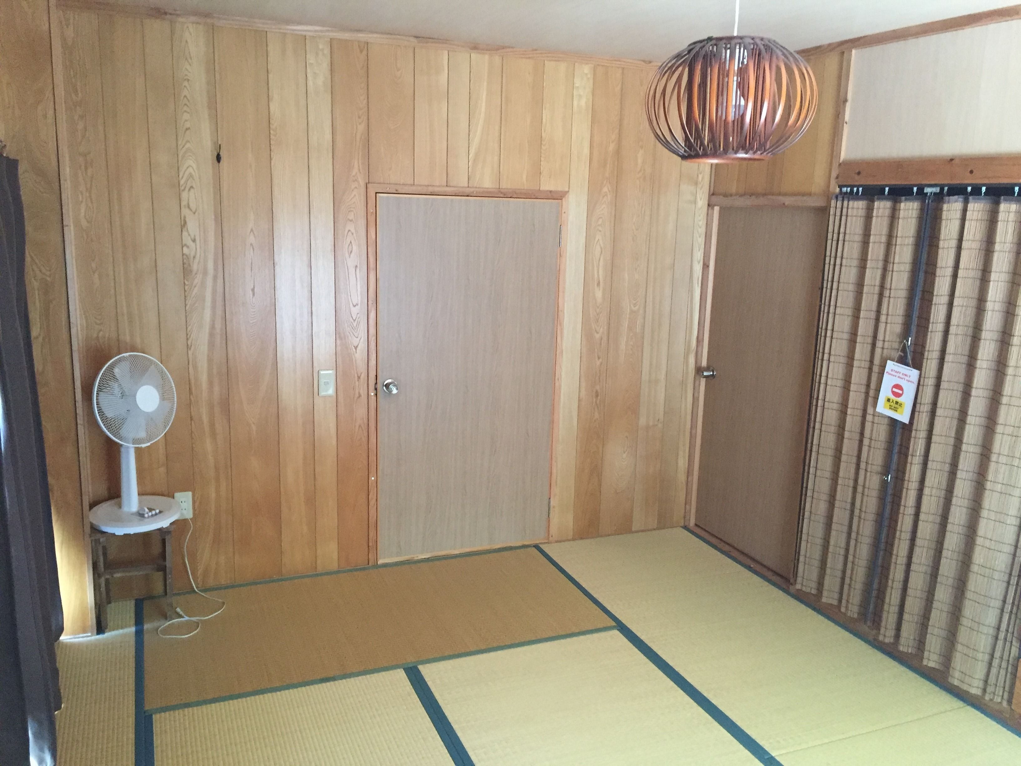 Une pièce aux murs recouverts de lambris en bois, dotée d’un sol en tatamis, d’un ventilateur sur pied et d’une porte sur laquelle est apposée une pancarte.