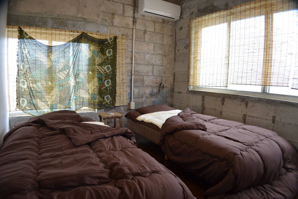 A room with two beds, a small table, and a window with blinds.