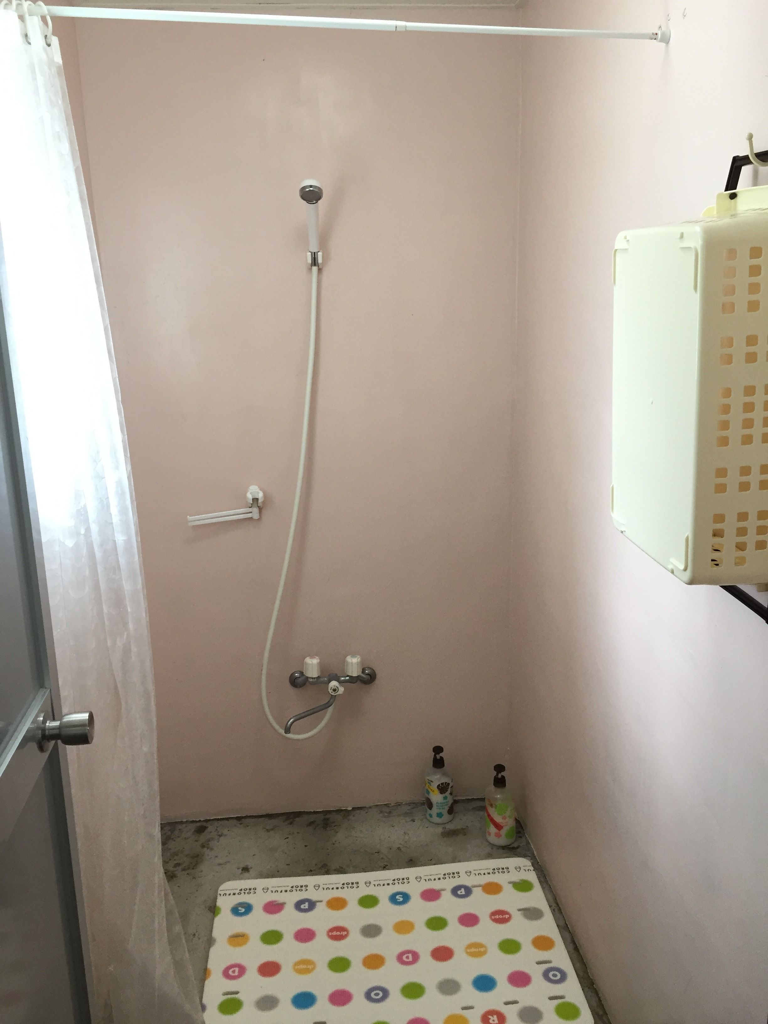 A small, enclosed shower area with a white showerhead and control knobs, a white plastic container on the wall, and a colorful mat on the floor.