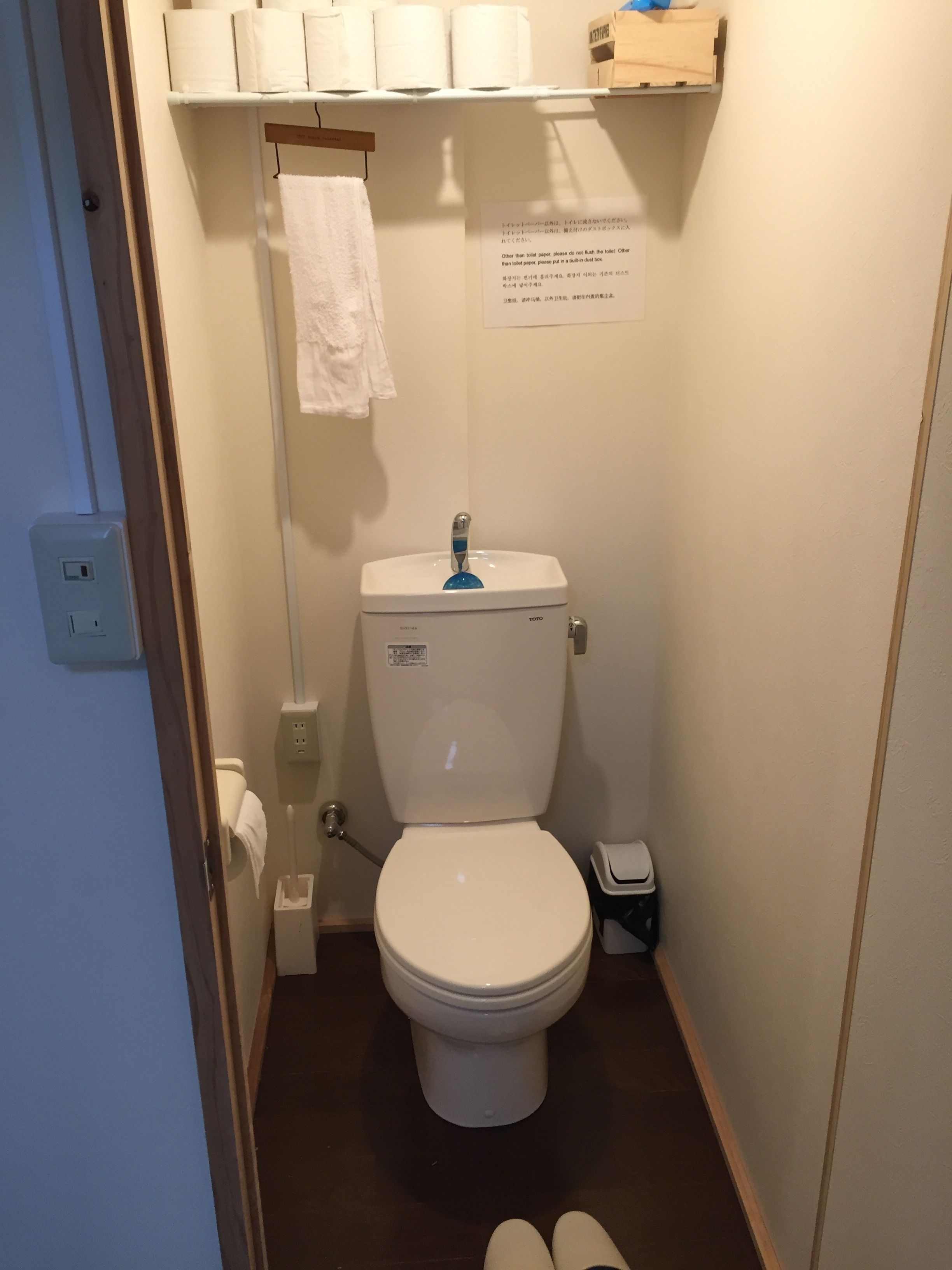 A small, compact bathroom with a white toilet, a blue flush handle, and a white towel hanging on a rack. There is a sign with instructions on the wall, and a small trash bin is visible to the right of the toilet.