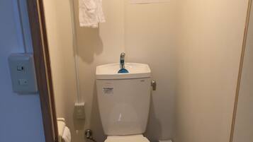 A small, compact bathroom with a white toilet, a blue flush handle, and a white towel hanging on a rack. There is a sign with instructions on the wall, and a small trash bin is visible to the right of the toilet.