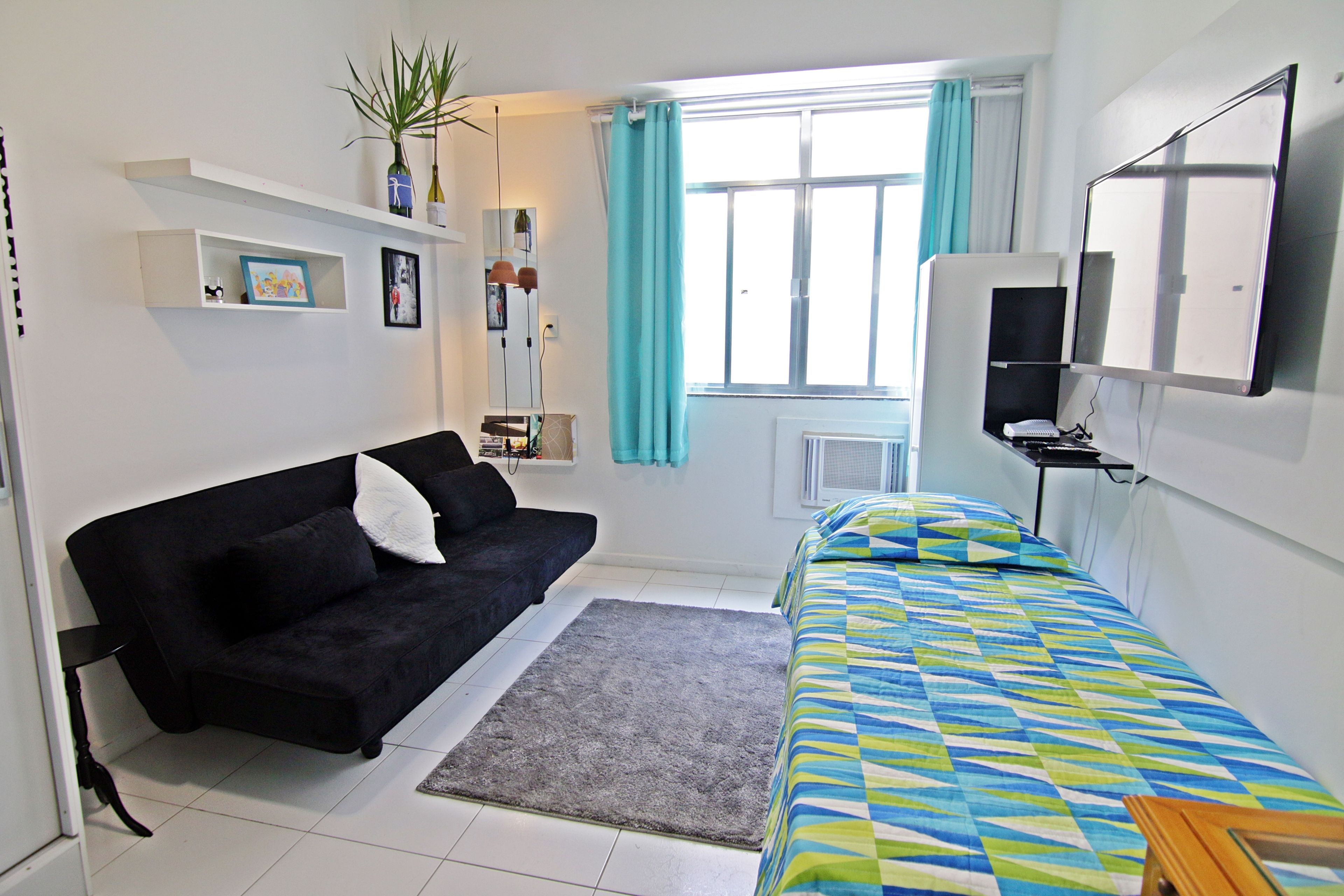 A room with a black sofa, a bed with a colorful quilt, a TV on a stand, a window with curtains, and a plant on a shelf.