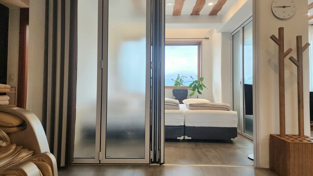 A modern hotel room with a sliding glass door leading to a balcony, a bed with a view, and a wooden decor piece on the wall. - Palm Tree Villa (Seogwipo)