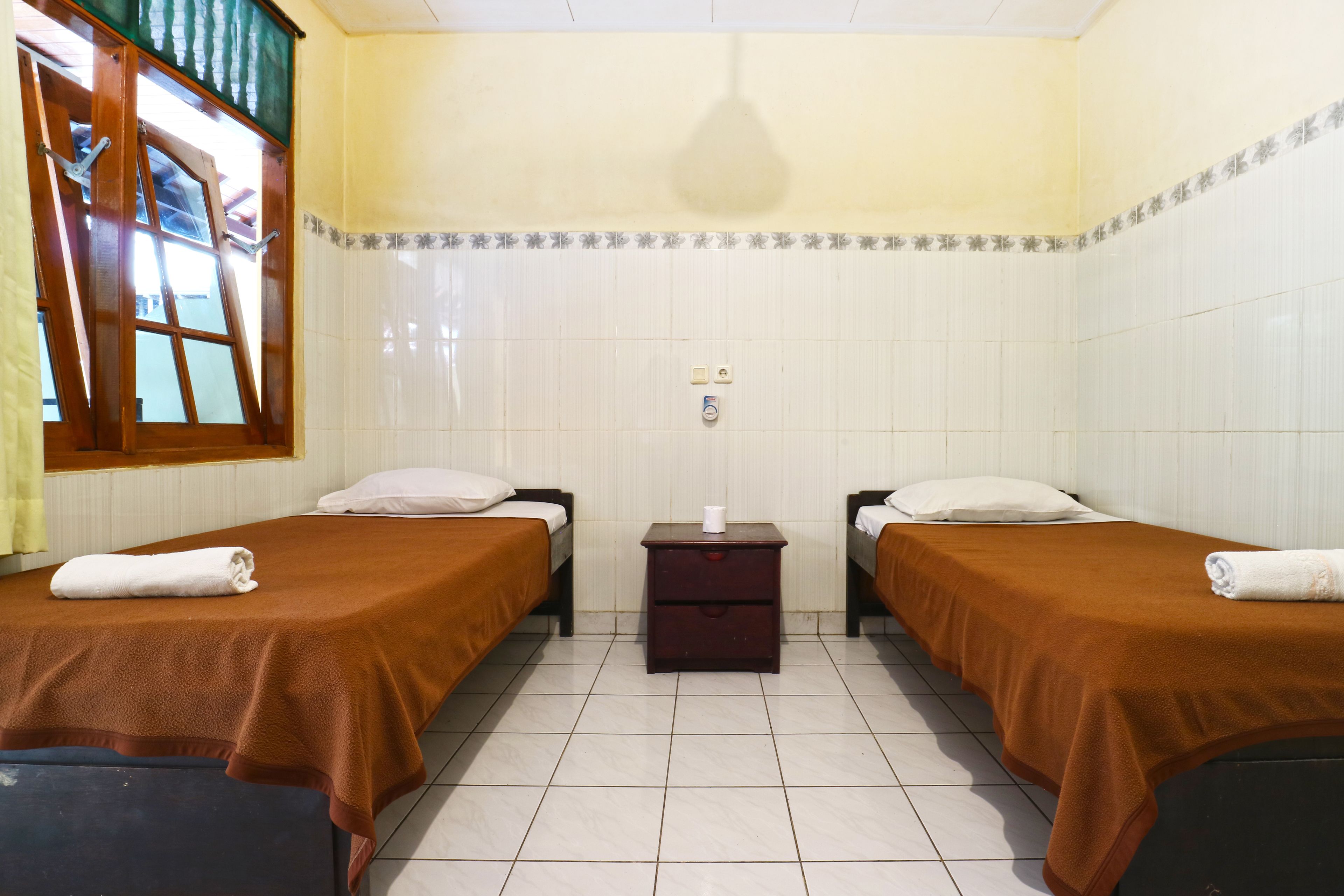 A room with two beds, a small wooden nightstand, and a window with a curtain.