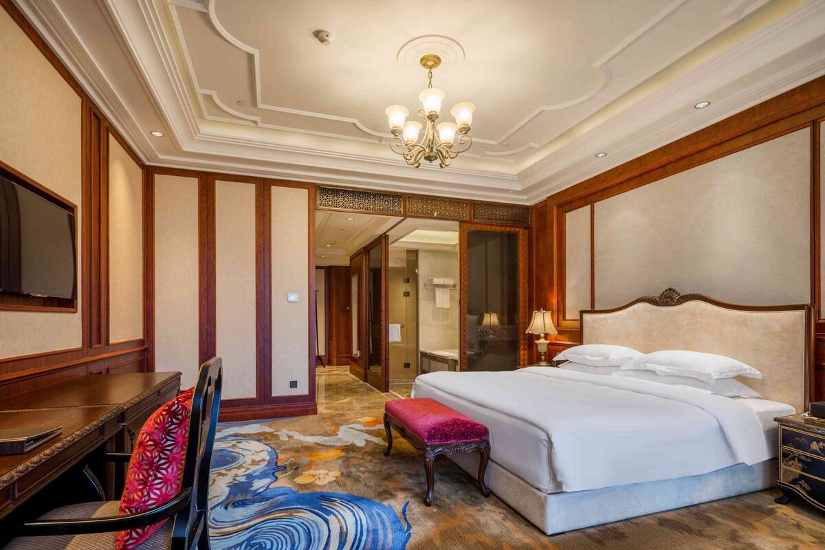 A hotel room with a bed, a desk, a chair, a television, and a bathroom visible through an open door.