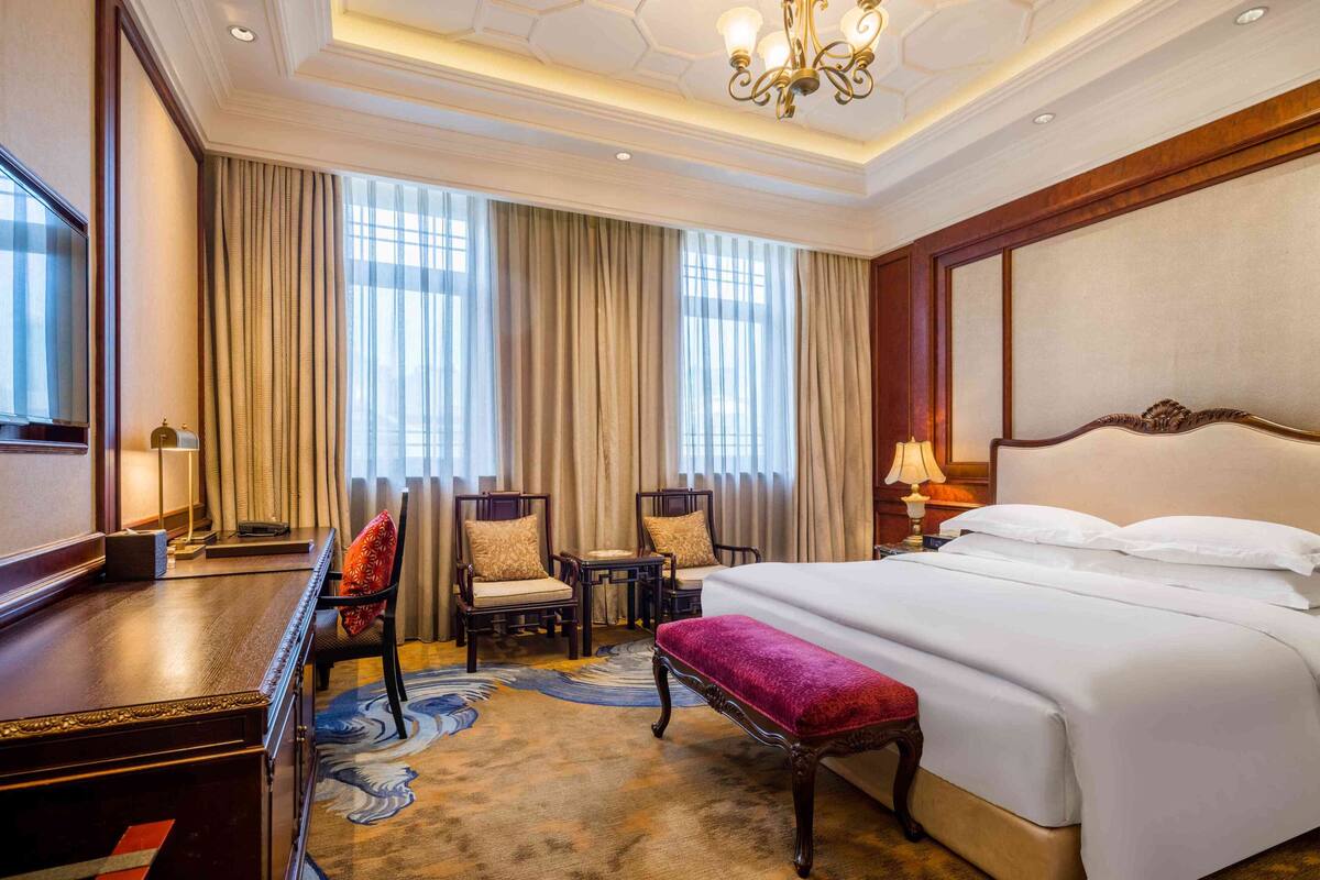A hotel room with a large bed, a desk, two chairs, a small table, and a bench.
