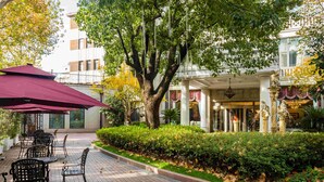 An outdoor patio area with tables and chairs, a large umbrella, and a building with columns and a fountain in the background. - Shanghai Donghu Hotel (Shanghai)