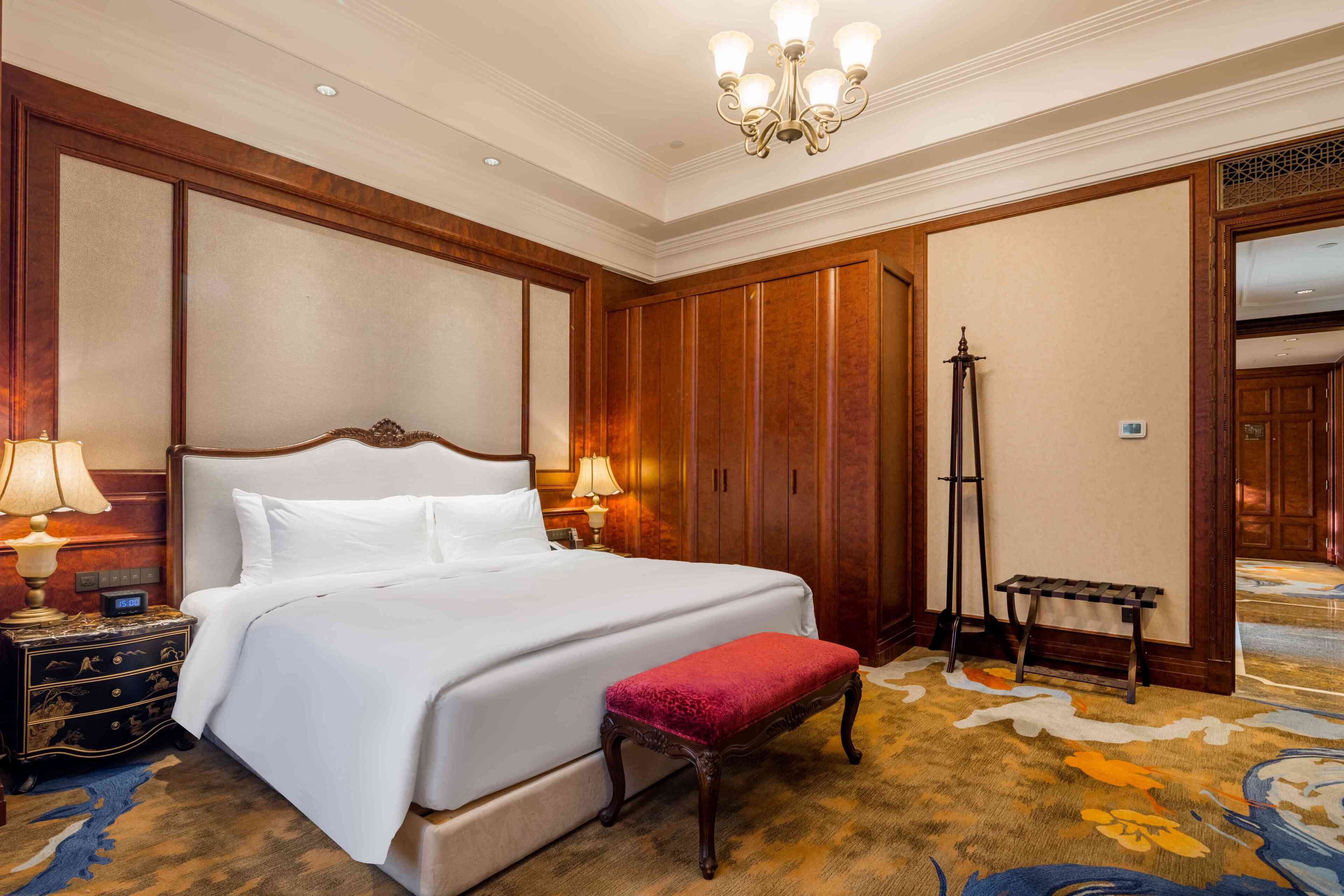 A hotel room with a large bed, wooden paneling, a standing lamp, a bench, and a carpeted floor.