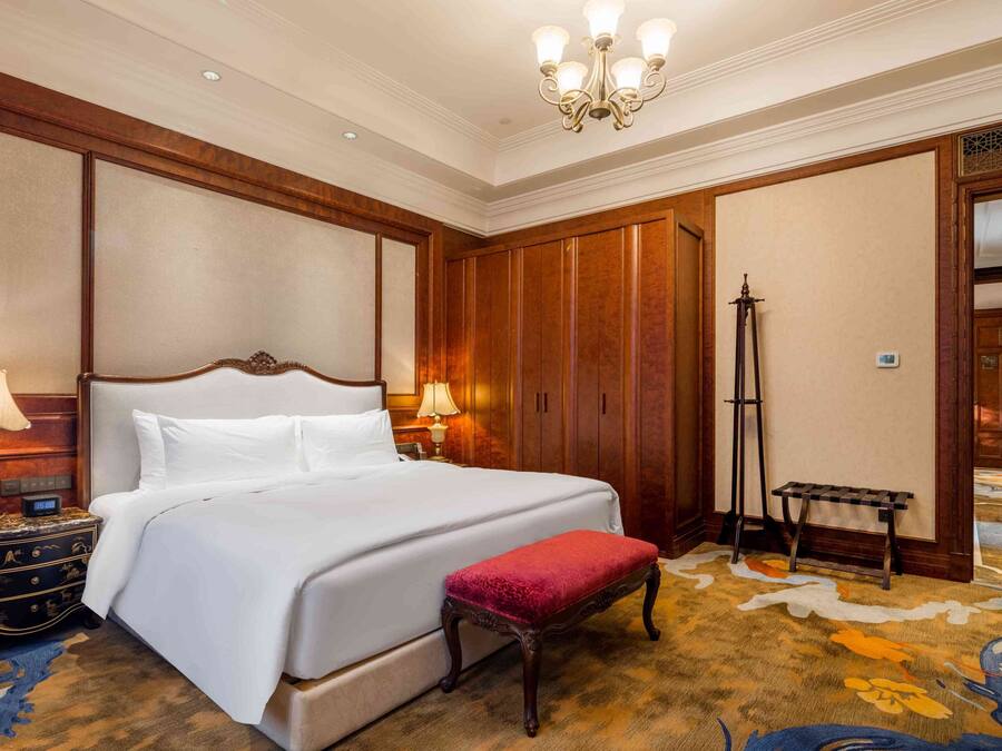 A hotel room with a large bed, wooden paneling, a standing lamp, a bench, and a carpeted floor.