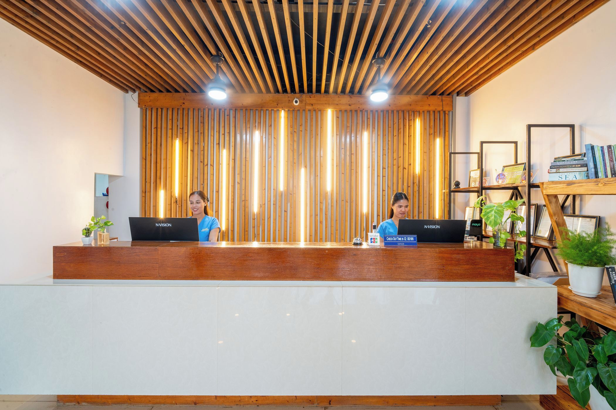 A reception desk with two staff members, a wooden slatted ceiling, and a bookshelf with books and decorative items.