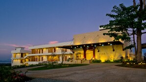 Un hotel de varios pisos con diseño moderno, que presenta una entrada destacada con un corredor cubierto y un árbol grande a la derecha.
