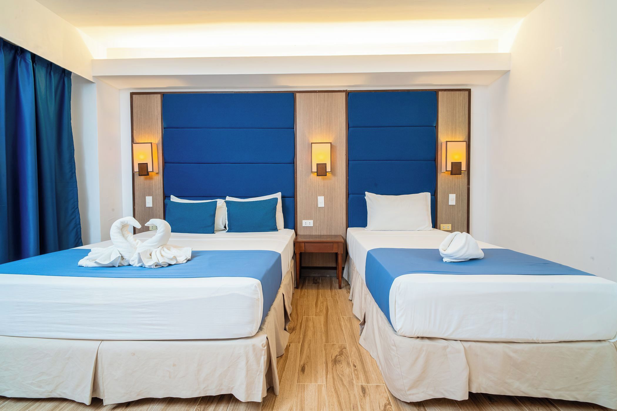A hotel room with two beds, each with a blue headboard and white bedding, and a wooden nightstand between them.