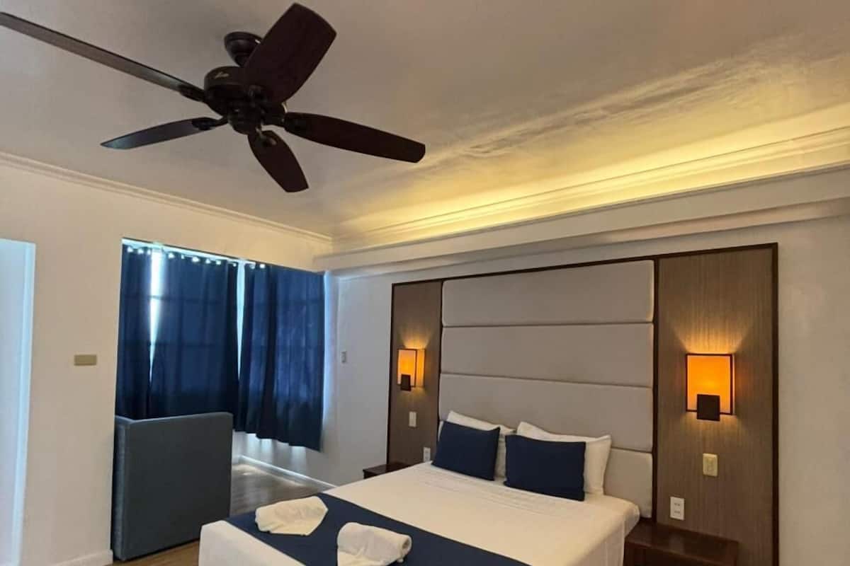Una habitación de hotel con cama, ventilador de techo, mesita de noche y ventana con cortinas.
