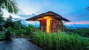 A wooden cabin with a thatched roof surrounded by lush greenery, accessible via a wooden deck.