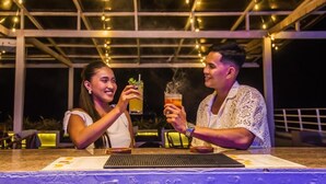 Dos personas brindando con bebidas en un bar con techo de vidrio y guirnaldas de luces.