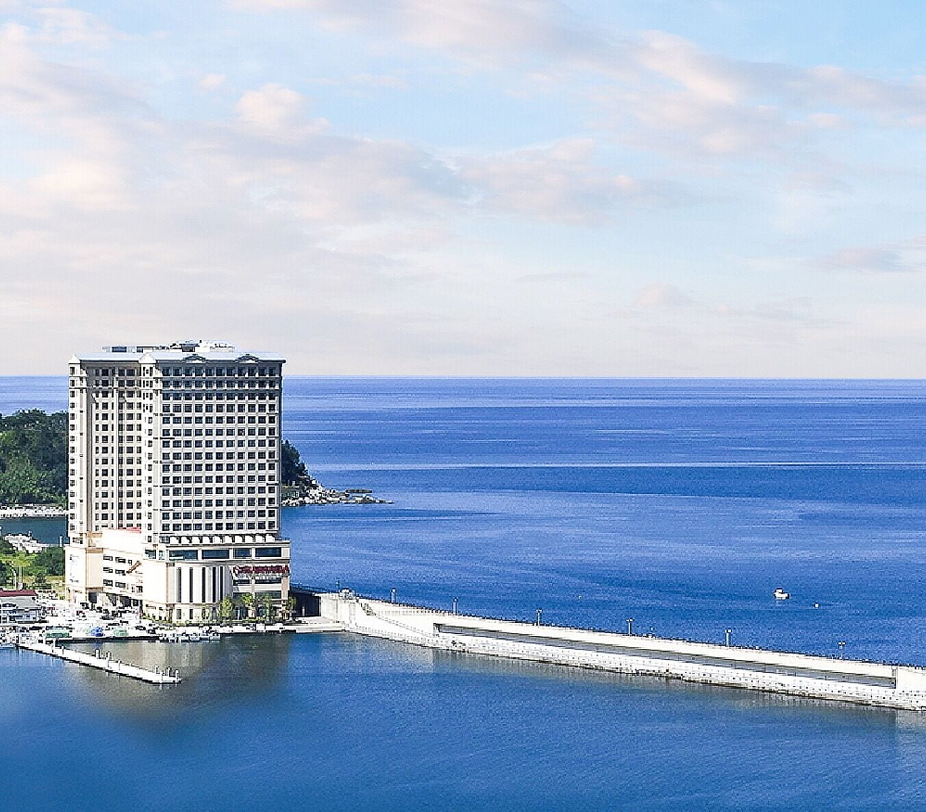 A high-rise hotel with a waterfront view, a long pier extending into the water, and a clear sky.