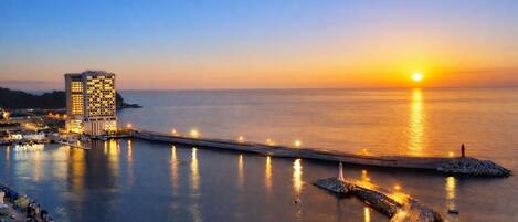 A coastal cityscape at sunset with a prominent hotel, a pier extending into the water, and a lighthouse.