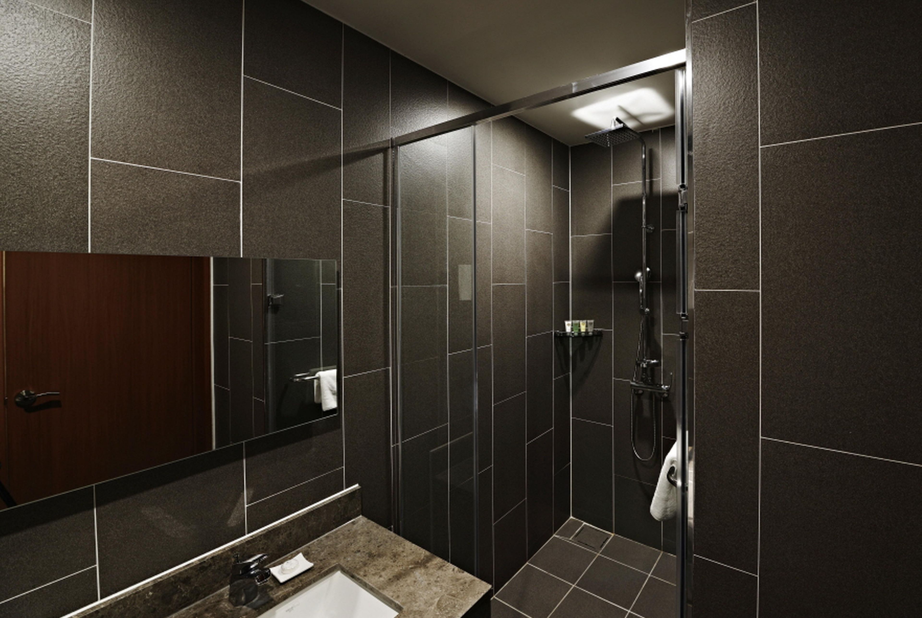 A modern bathroom with a large mirror, dark tiles, a shower, and a sink.