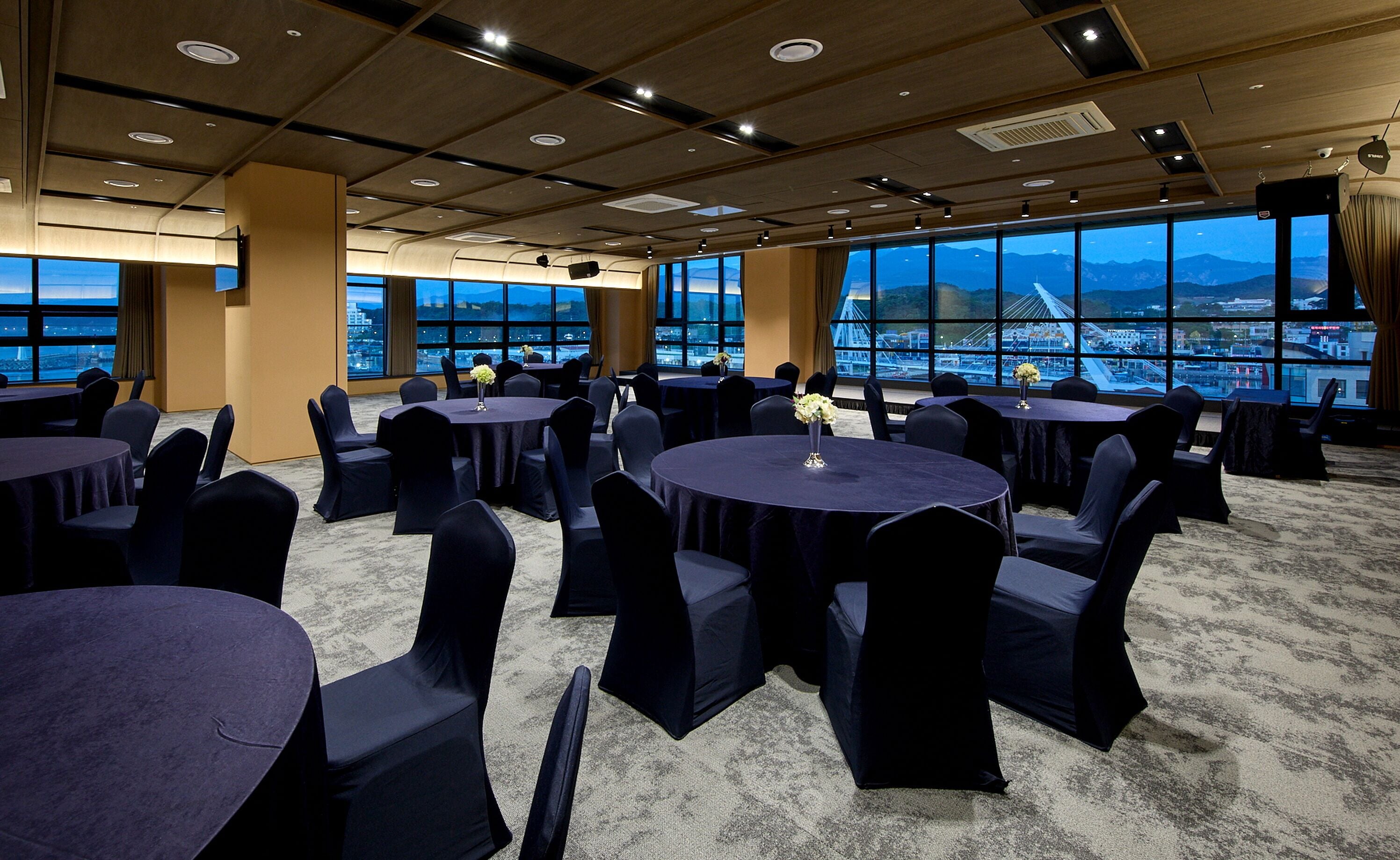 A conference room with multiple tables and chairs, large windows with a cityscape view, and a modern ceiling design.