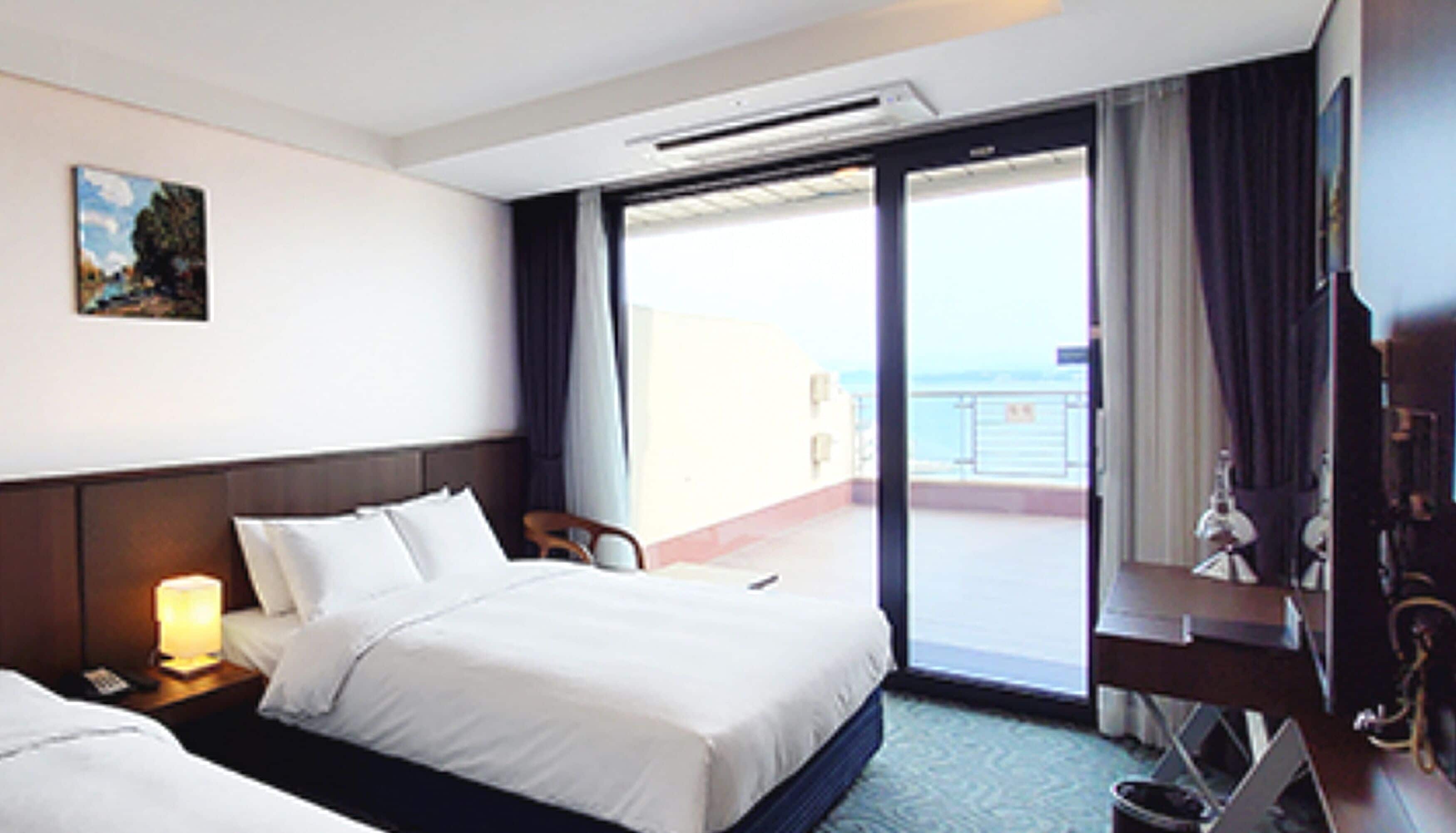 Habitación de hotel con cama, escritorio, silla, lámpara y vista al mar a través de una puerta corrediza.