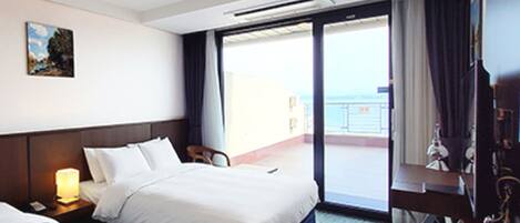 Habitación de hotel con cama, escritorio, silla, lámpara y vista al mar a través de una puerta corrediza.