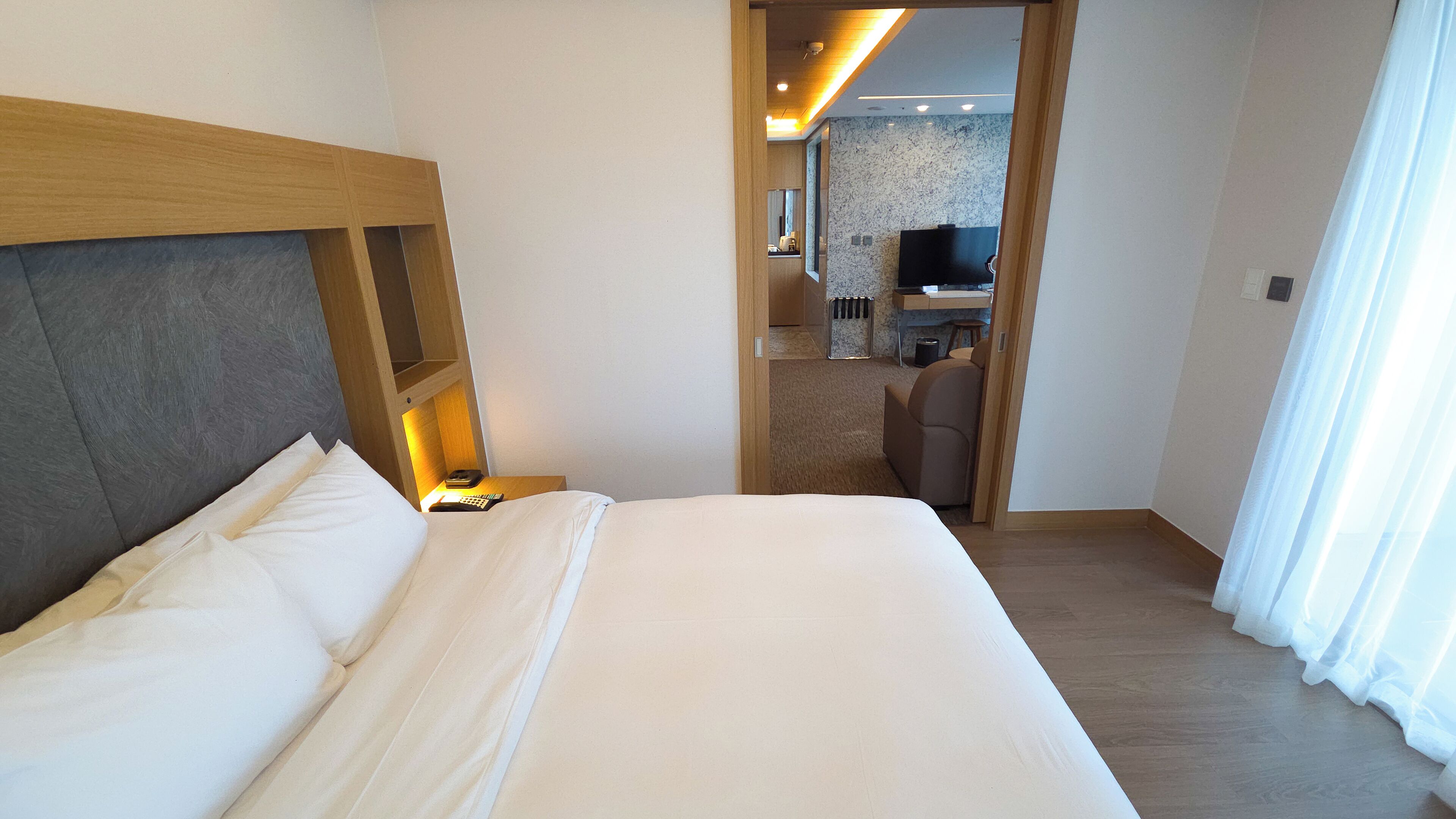 A hotel room with a large bed, a wooden headboard, a bedside table with a lamp, and a view into the room's kitchen area.