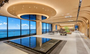 A spacious indoor area with large windows offering a view of the ocean, a central pool, and a seating area with tables and chairs.