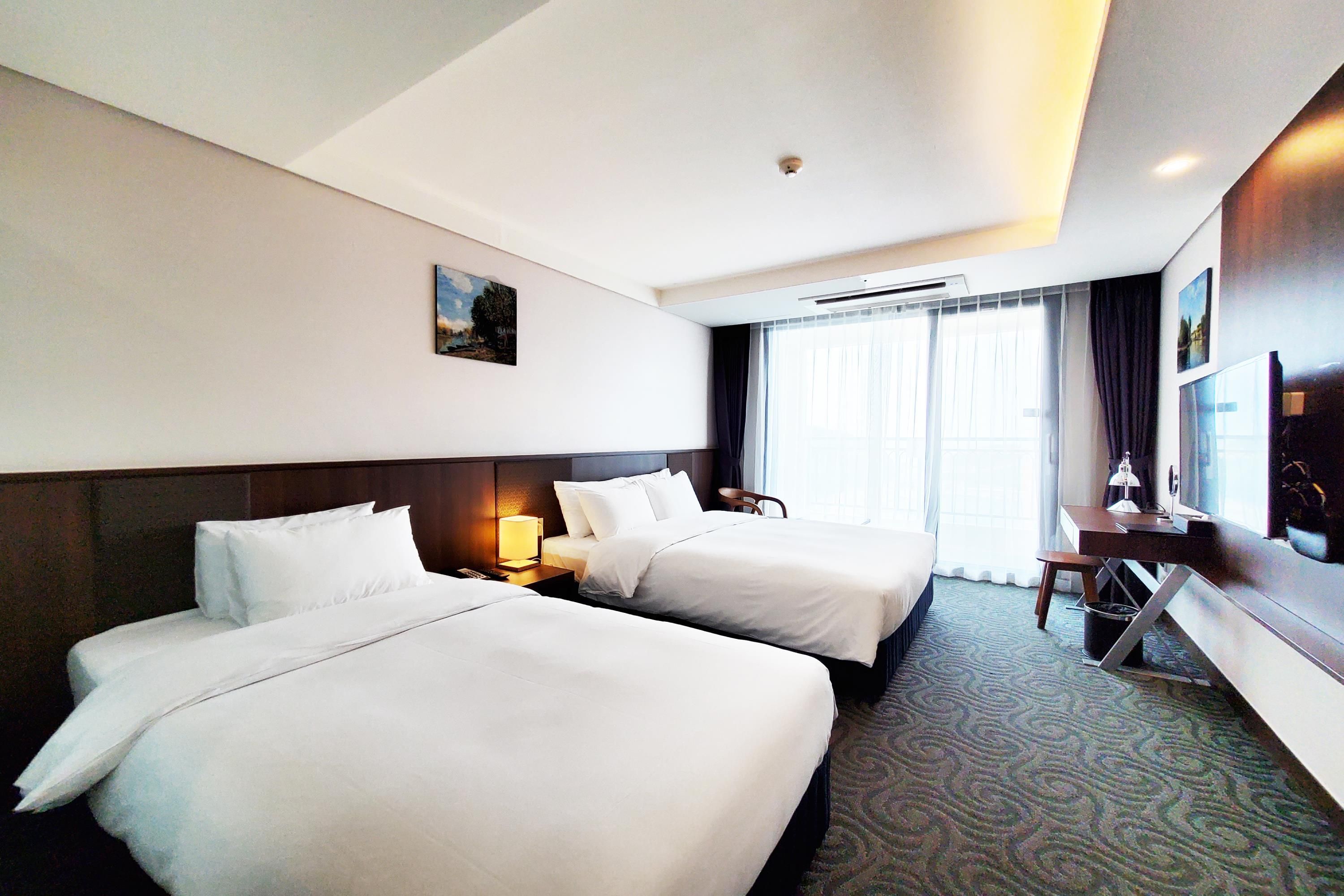 A hotel room with two beds, a desk, a TV, and a window with curtains.