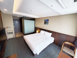 Habitación de hotel con una cama grande, una silla de madera, una mesita de noche con una lámpara, un cuadro en la pared y una puerta que da a otra habitación.