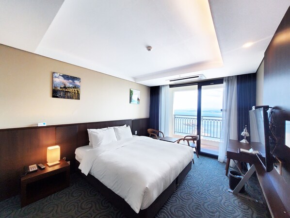 A hotel room with a large bed, a desk with a lamp, a TV, and a view of the sea through the balcony.