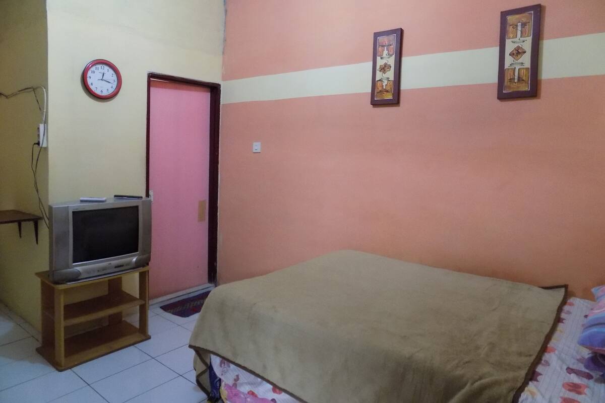 Una habitación con una cama, un televisor sobre un mueble, un reloj y dos cuadros en la pared.