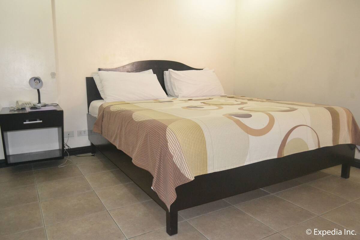 Een net opgemaakt bed met een bedsprei met een patroon, een nachtkastje met een lamp, en een stopcontact op de muur.