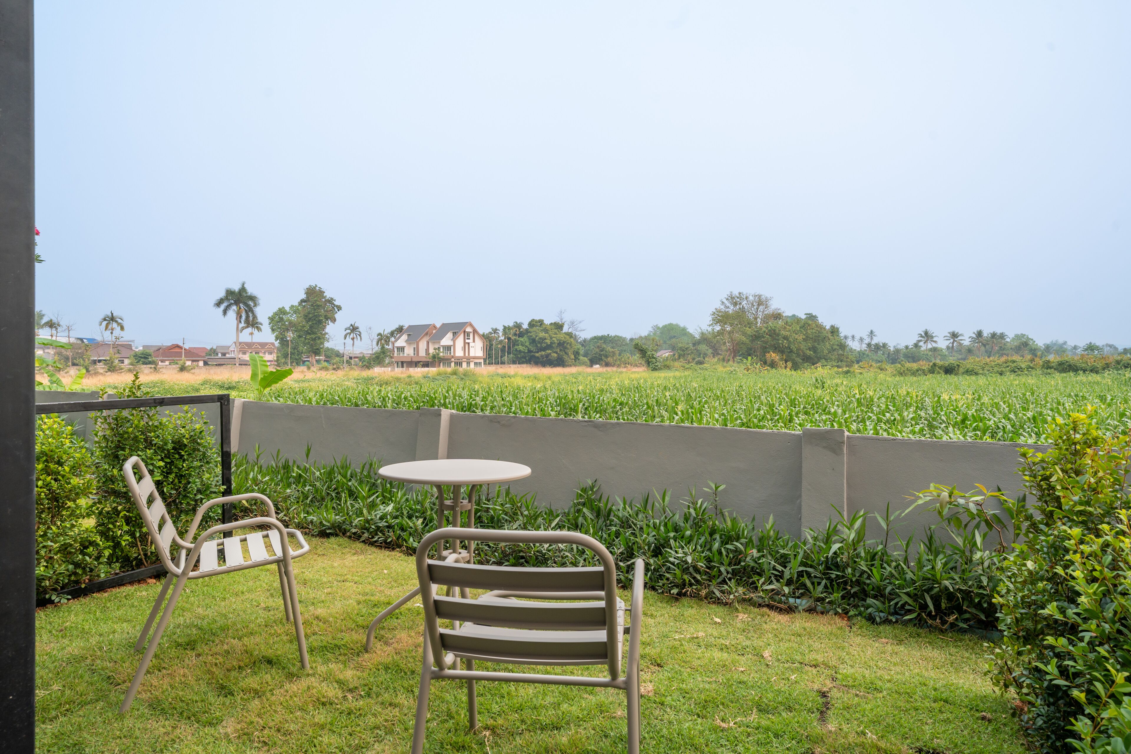 A patio with two metal chairs and a small round table, surrounded by a low concrete wall, overlooking a field and distant buildings.