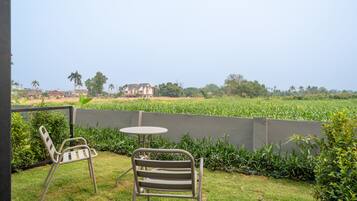 A patio with two metal chairs and a small round table, surrounded by a low concrete wall, overlooking a field and distant buildings.