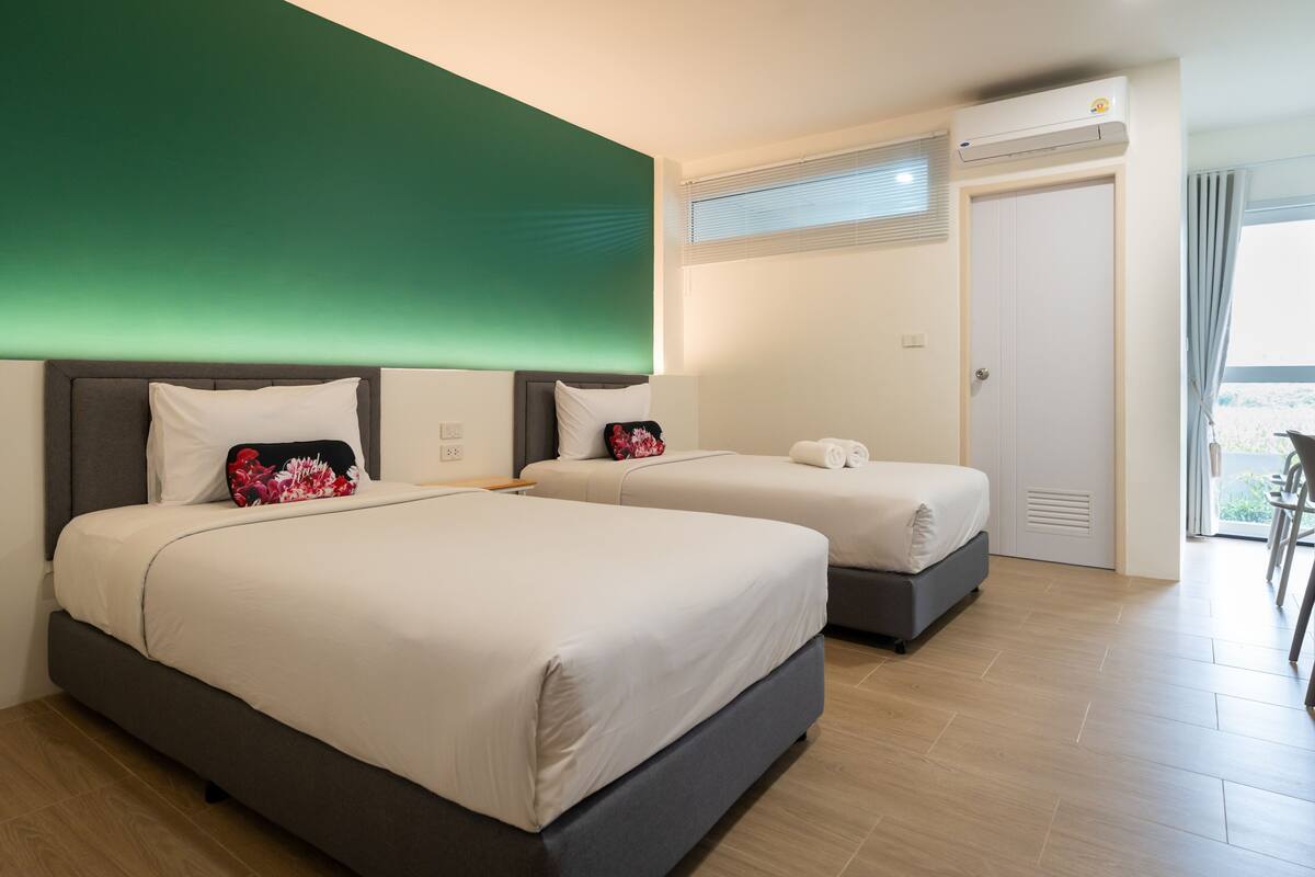 A hotel room with two beds, a green accent wall, an air conditioning unit, and a view of the outdoors.