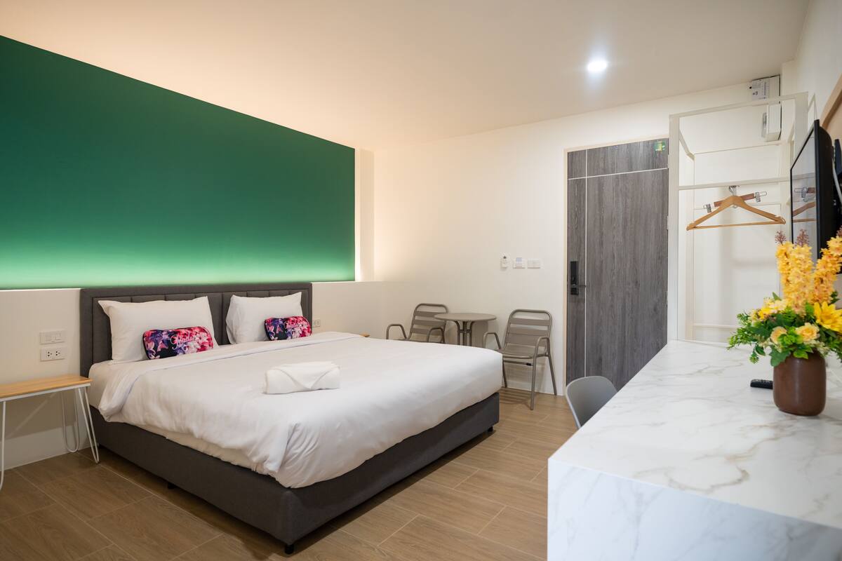A hotel room with a bed, bedside table, chair, desk, and a bathroom with a mirror and towel rack.