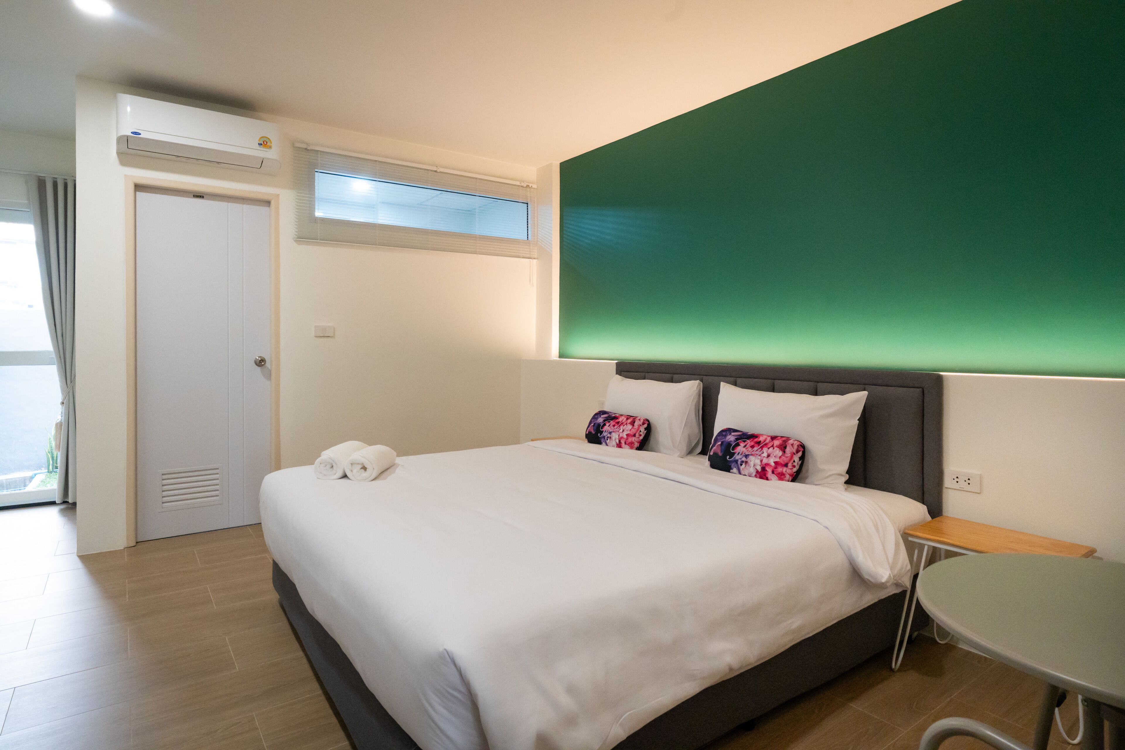 A hotel room with a bed, a small table, a green accent wall, a window with blinds, and an air conditioning unit.
