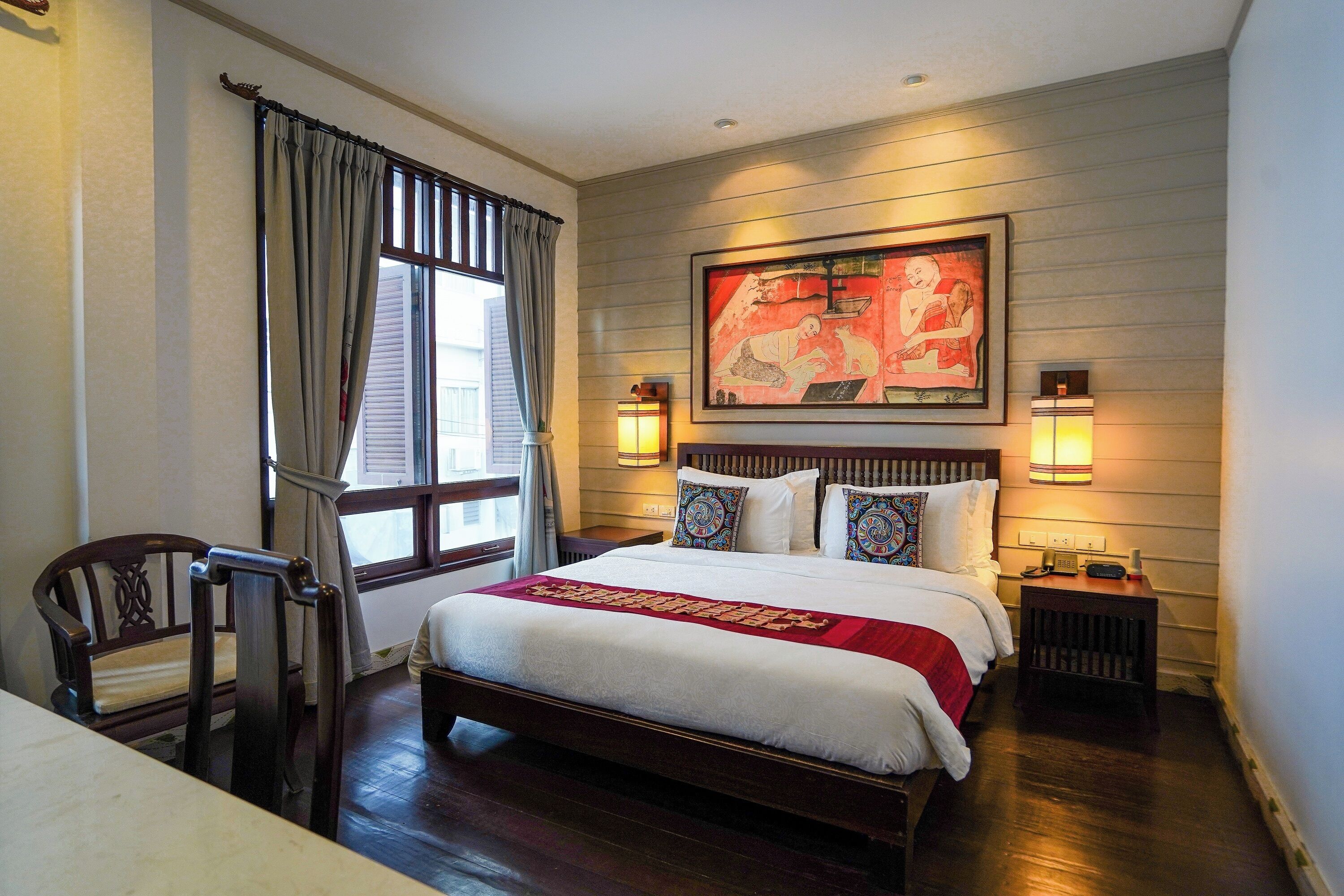 A hotel room with a large bed, a wooden headboard, a window with curtains, a wall-mounted painting, and two bedside lamps.