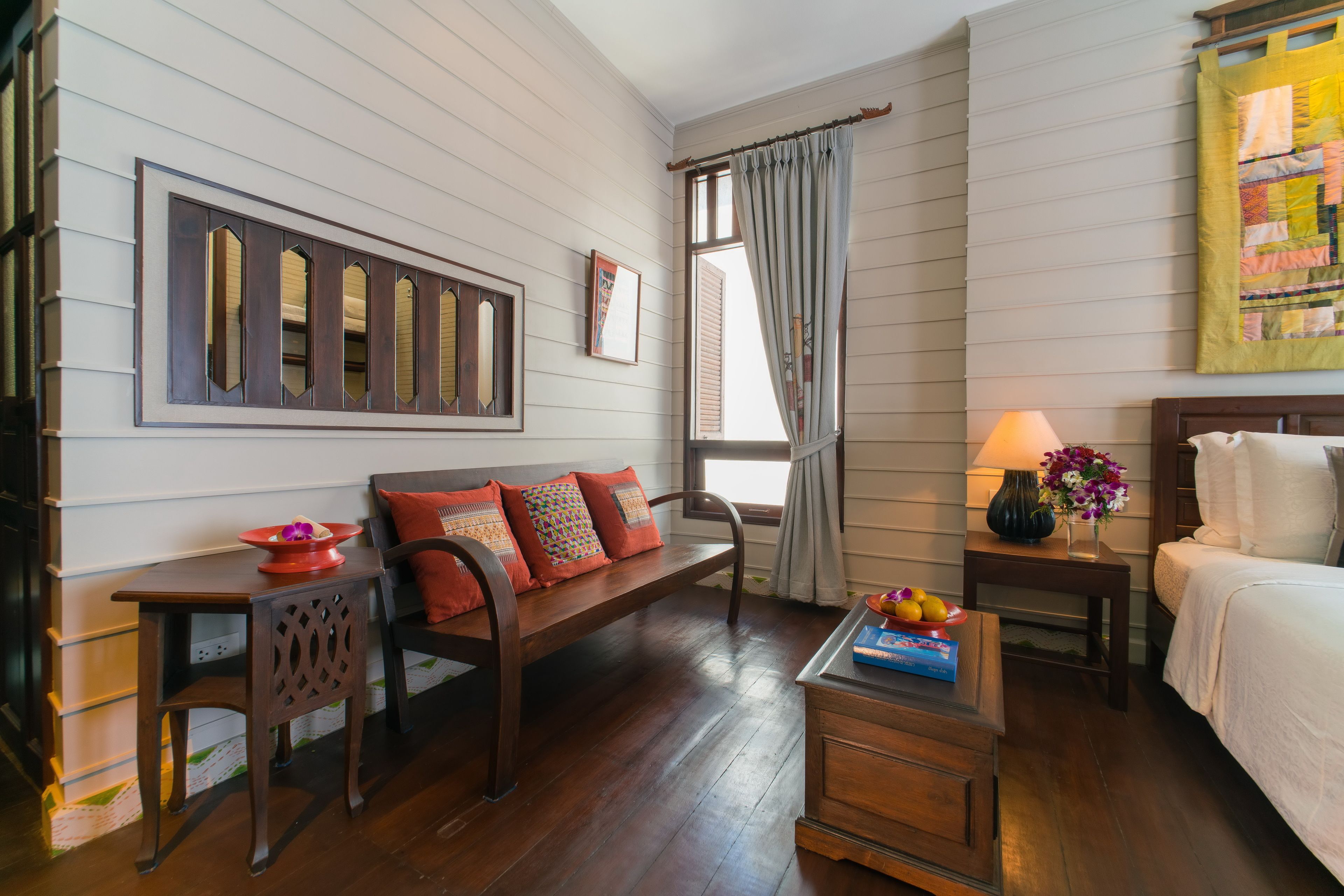 Una habitación con un banco de madera, una mesita con un jarrón y flores, una cama y una ventana con cortinas.