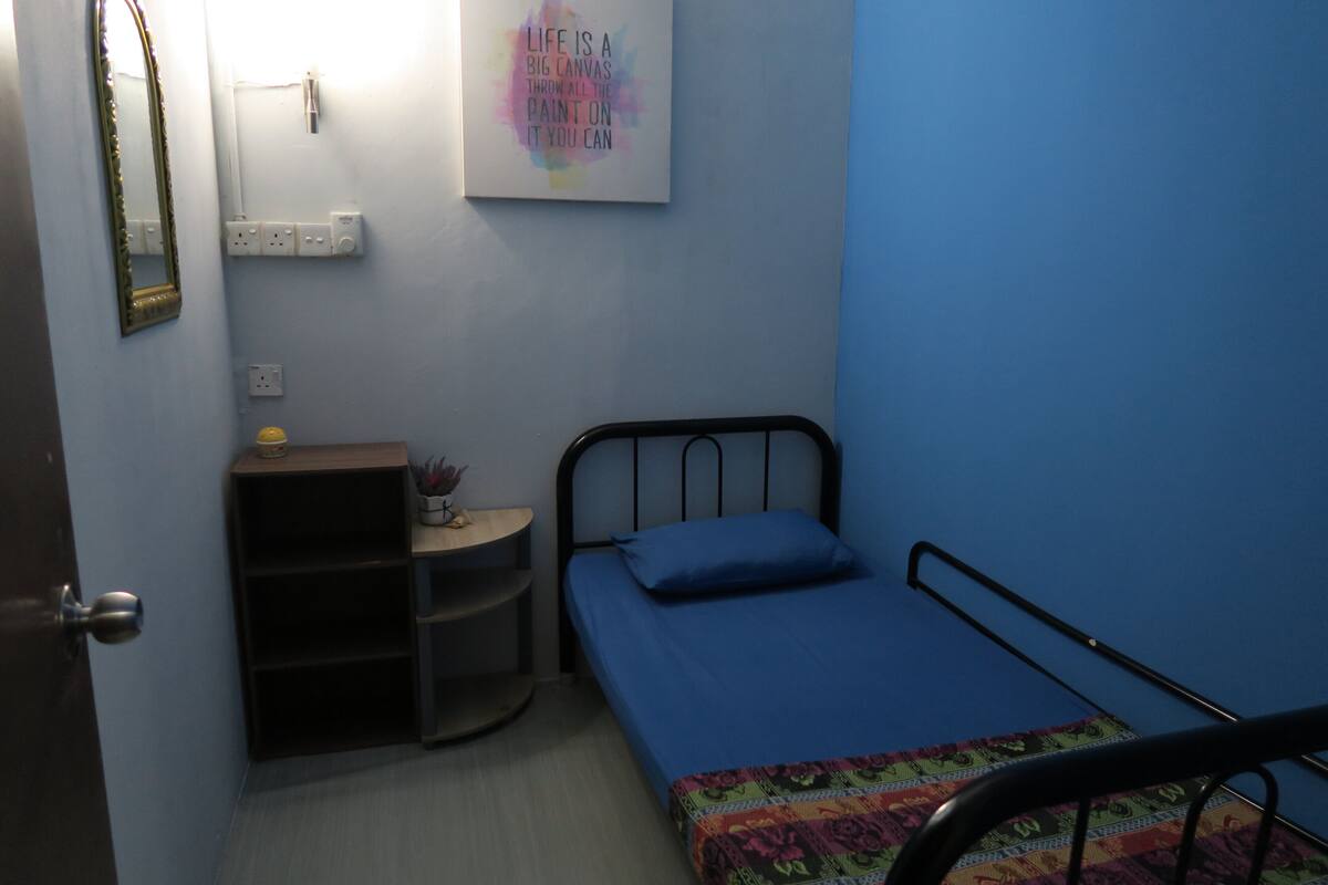 Ein kleines Zimmer mit einem Einzelbett, einer blauen Wand, einem Nachttisch mit Lampe und einem Wandspiegel.