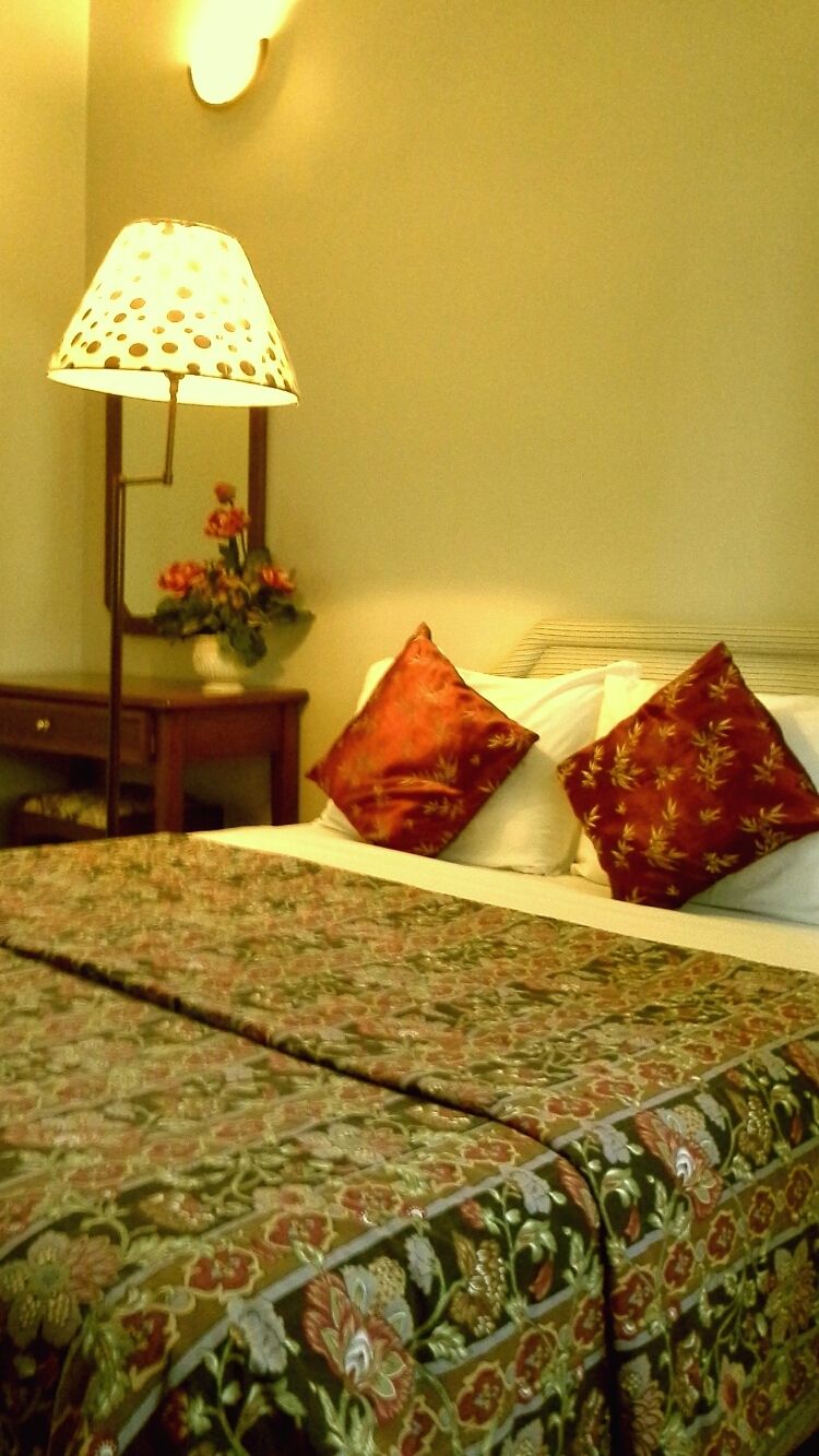 Una cama con colcha estampada, dos almohadas, una mesita de noche con lámpara y un florero con flores.