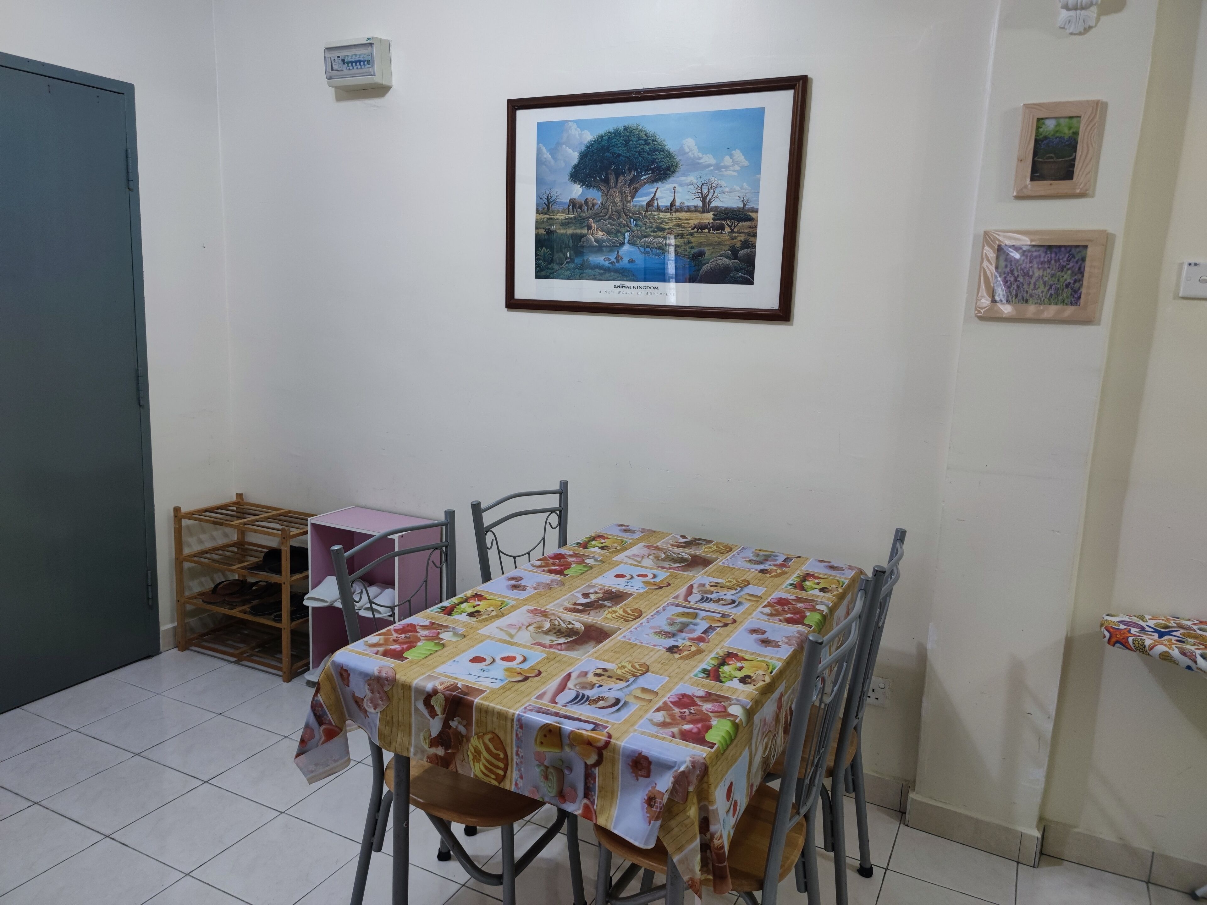 A dining area with a table covered in a patterned tablecloth, metal chairs, a pink storage unit, and a framed picture of a landscape on the wall.