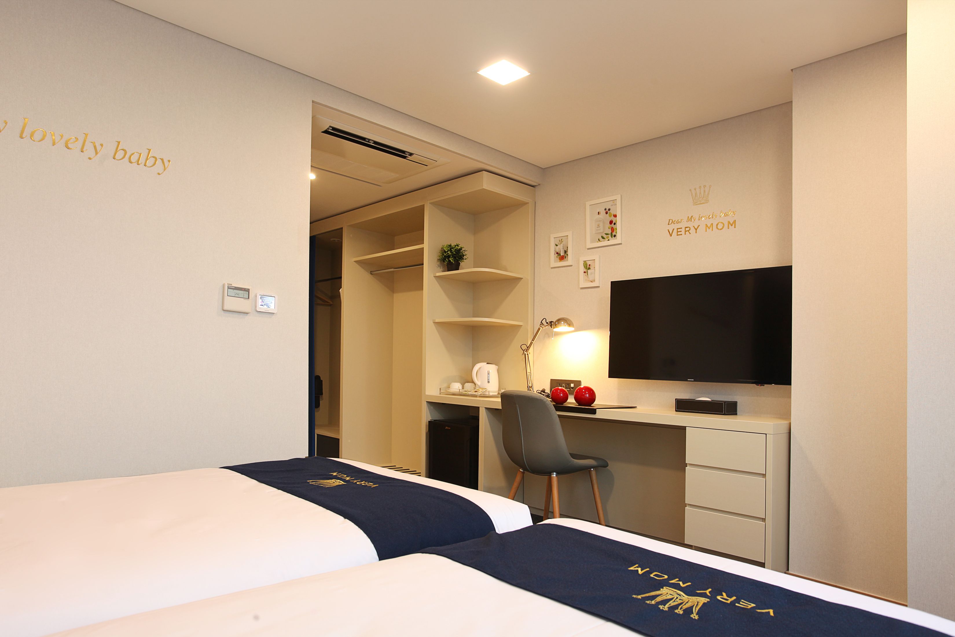 A hotel room with two beds, a desk with a computer, a TV, and a small kitchenette.