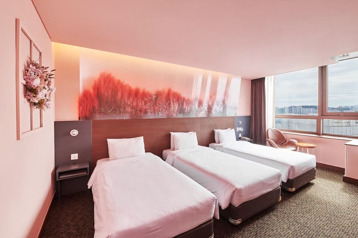 Hotellihuone, jossa on kolme erillistä sänkyä, suuri ikkuna, josta on näkymä kaupunkiin, ja seinällä moderni abstrakti maalaus.