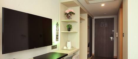 Una habitación de hotel moderna con una televisión de pantalla plana grande, un escritorio con una silla, una cafetera pequeña y una estantería con un jarrón de flores.