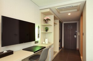 Una habitación de hotel moderna con una televisión de pantalla plana grande, un escritorio con una silla, una cafetera pequeña y una estantería con un jarrón de flores.