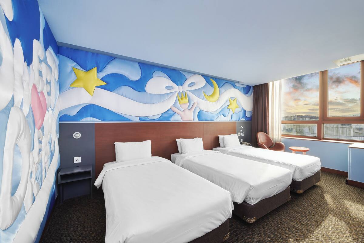 Hotellihuone, jossa on kaksi sänkyä, seinällä suuri seinämaalaus, ikkuna, josta on näkymä kaupunkiin, ja pieni pöytä.