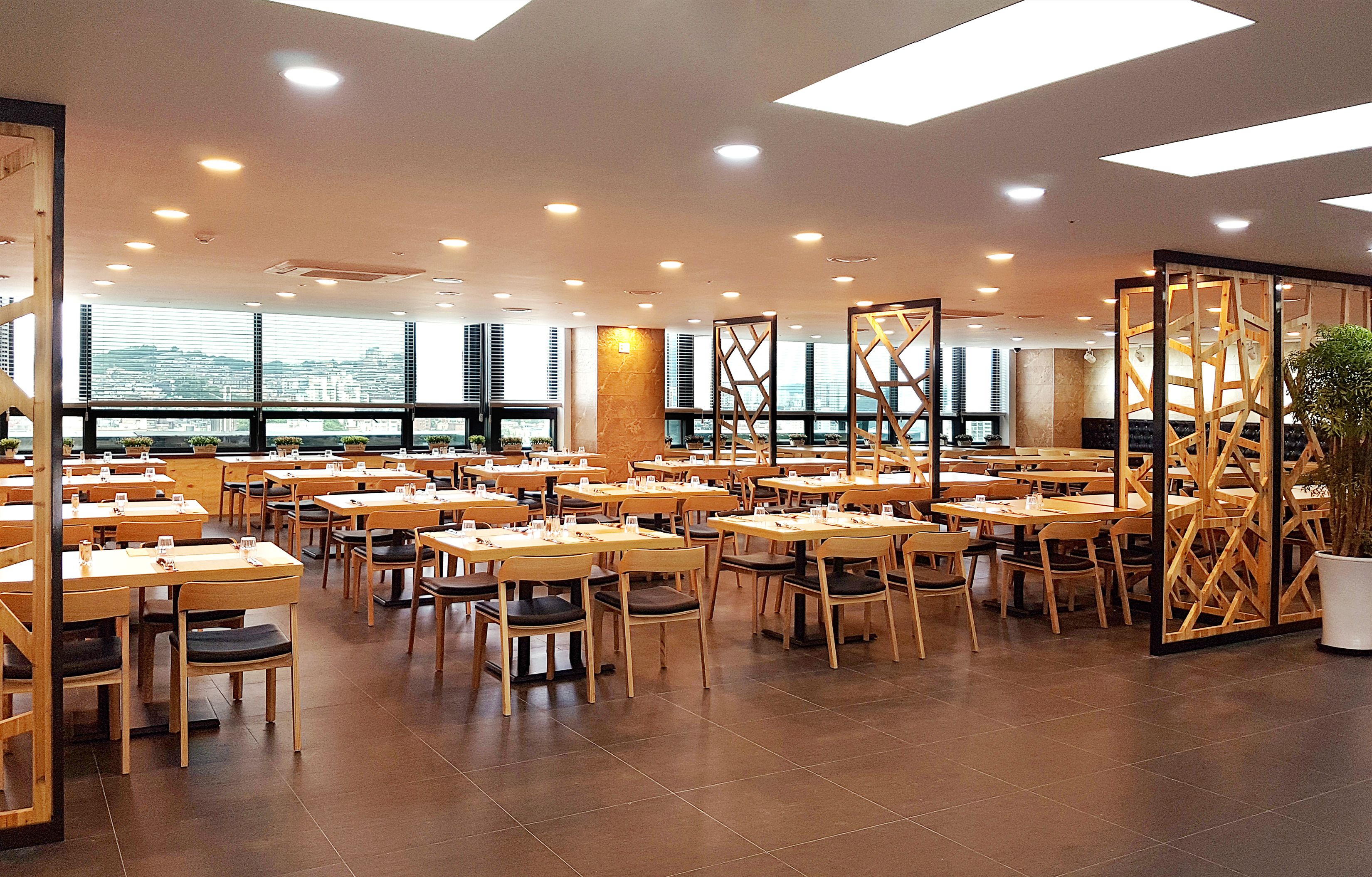 Un comedor amplio con varias mesas y sillas, ventanas grandes que ofrecen vista a la ciudad y un diseño de techo moderno.